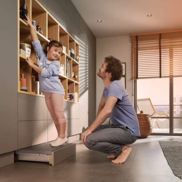 A father and daughter in front of a wall unit. The daughter is standing on a SPACE STEP plinth solution and reaches the top shelf.