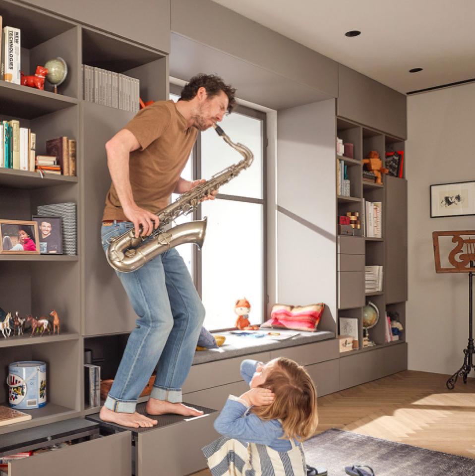 A man plays saxophone while standing on the SPACE STEP plinth step by Blum. His daughter covers her ears as she watches him.