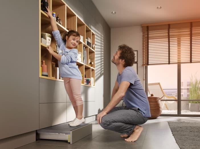 A father and daughter in front of a wall unit. The daughter is standing on a SPACE STEP plinth solution and reaches the top shelf.