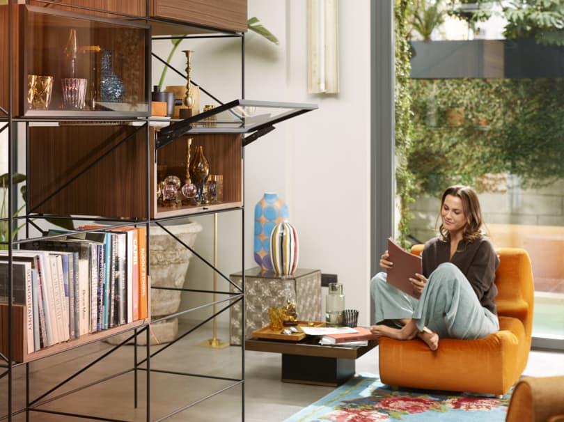 A woman in a modern living space. In the avant-garde shelving unit, an open wall cabinet shows the hidden AVENTOS HKi lift system.